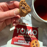 YOMO gluten free ready-made Choc Chip Gingerbread cookie held above its packaging with a cup of tea in the background. Gluten free and vegan cookie