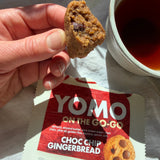 YOMO gluten free ready-made Choc Chip Gingerbread cookie held above its packaging with a cup of tea in the background. Gluten free and vegan cookie