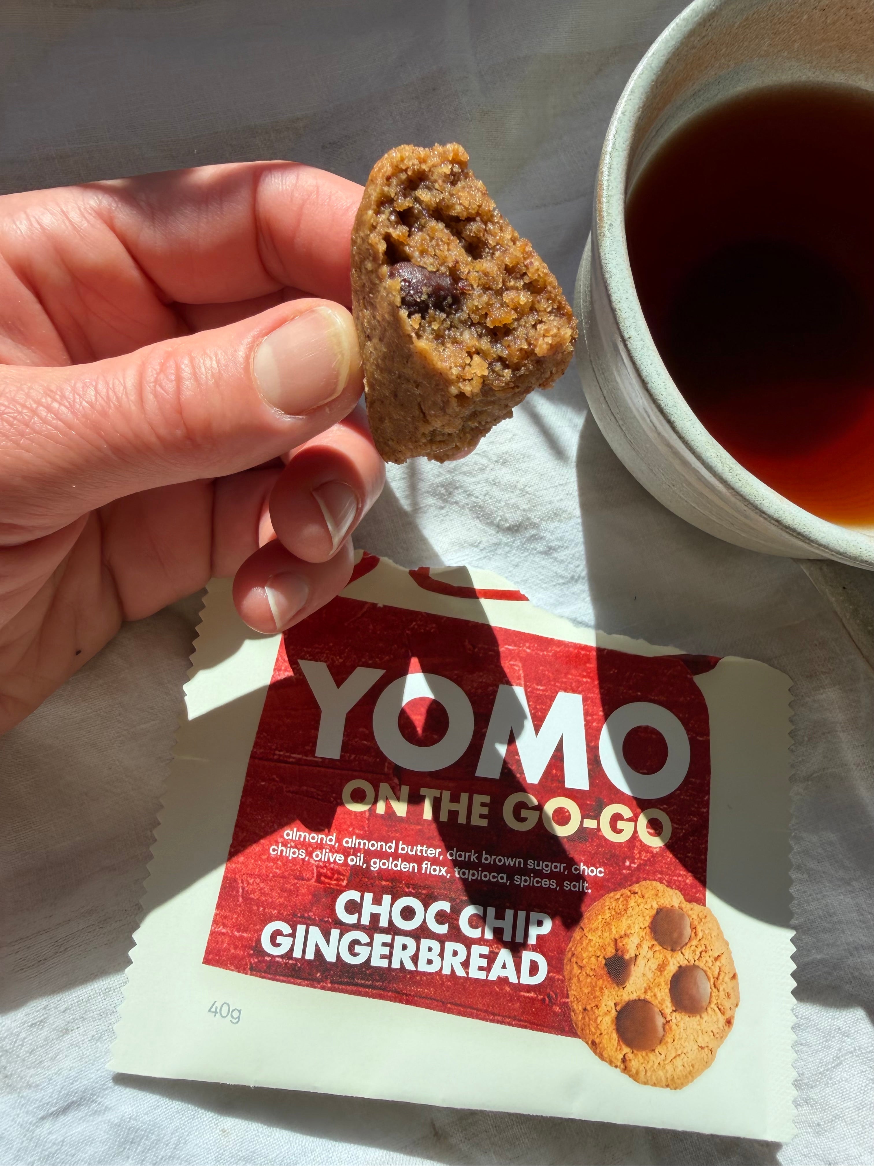 YOMO gluten free ready-made Choc Chip Gingerbread cookie held above its packaging with a cup of tea in the background. Gluten free and vegan cookie